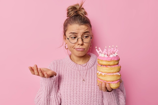 Free photo questioned sad beautiful woman raises palm looks displeased at delicious doughnuts with burning candles feels unhappy as celebrates birthday alone wears round spectacles and knitted sweater