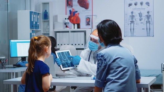 Radiologist explaining x-ray using tablet in medical office and nurse working on computer. Pediatrician specialist with protection mask providing health care service radiographic treatment examination