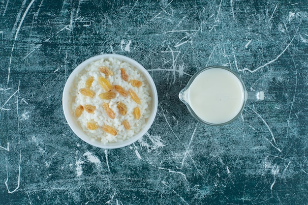 Raisins on a bowl of rice pudding next to a glass of milk, on the blue table. 