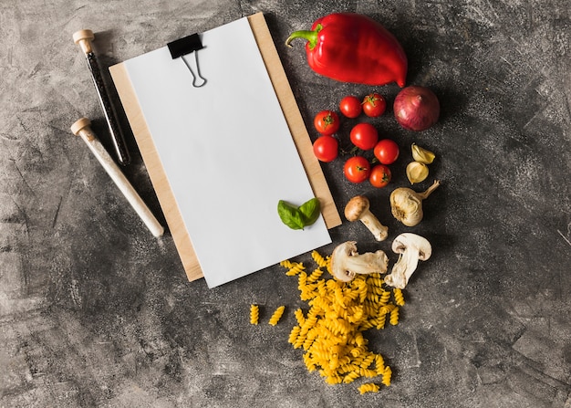 Free Photo raw fusilli with ingredients and paper on clipboard against grunge background