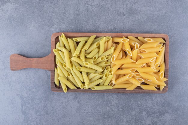 Raw green and yellow penne pasta on wooden board.