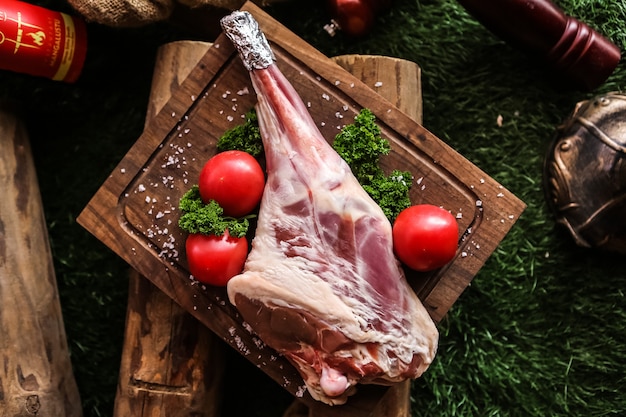 Raw lamb leg on the wooden board prepared for cooking salt tomato greens top view