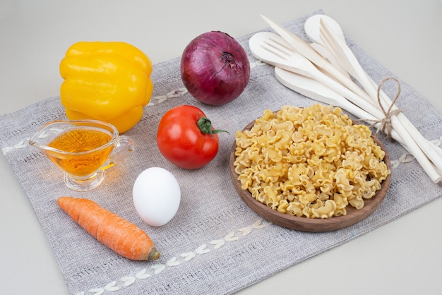 Raw macaroni with vegetables on wooden plate on tablecloth