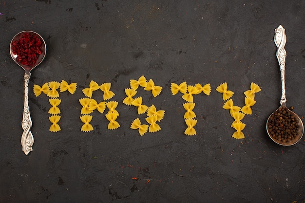 Free photo raw pasta tasty word shaped yellow along with two silver spoons with different spices on a dark desk