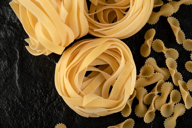 Raw tagliatelle nests and bow shaped pasta on black surface