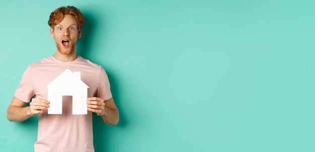 Free Photo real estate concept excited redhead guy with beard showing paper house cutout and gasping in awe sta