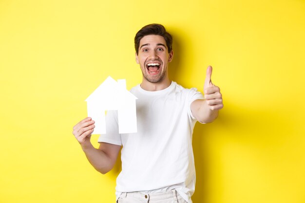 Real estate concept. Happy young male buyer showing thumb up and paper house model, smiling satisfied, standing over yellow background.