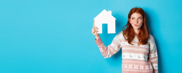 Free Photo real estate concept image of thoughtful redhead girl showing paper house model and thinking searchin