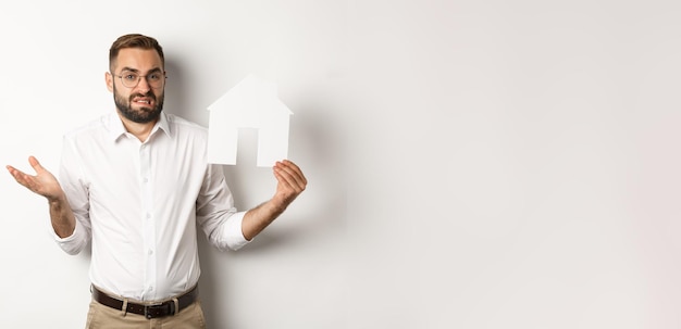 Free Photo real estate confused man shrugging showing house paper model and looking indecisive standing over wh