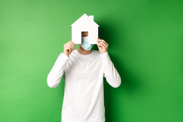 Free Photo real estate and covid-19 pandemic concept. funny young man in face mask and white long-sleeve hiding face behind paper house cutout, peeking at camera, green background