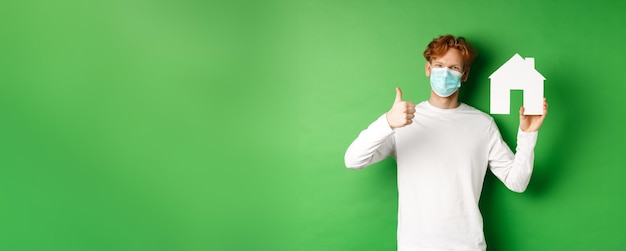 Free photo real estate and covid pandemic concept young redhead man in medical mask showing paper house cutout