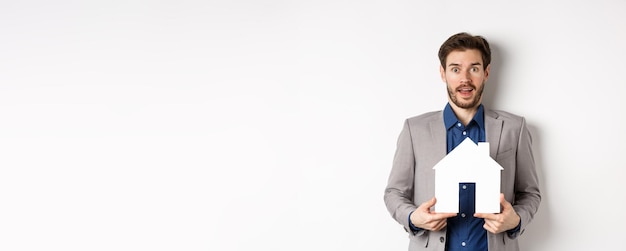 Free Photo real estate excited business man in suit holding paper house cutout and looking amazed at camera buy