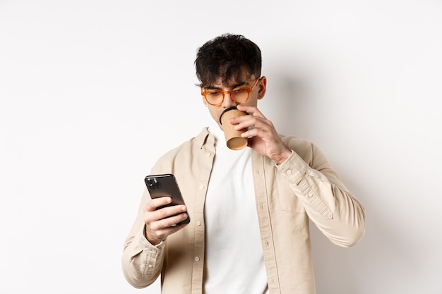 Real people. Young man reading smartphone screen and drinking coffee, looking at phone, standing on white background