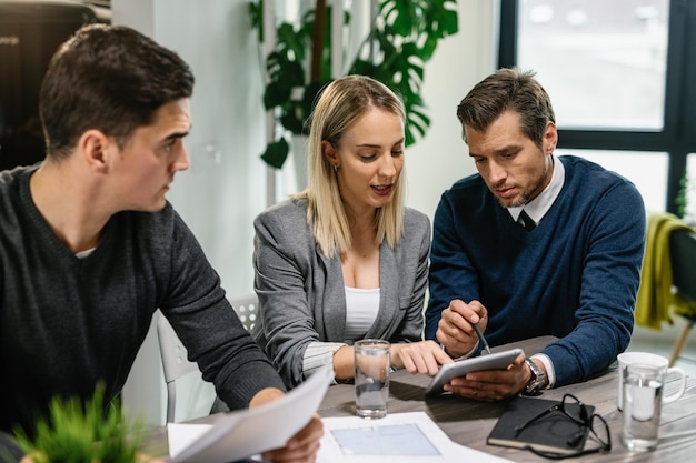 Rear estate agent and young couple going through housing plans on touchpad while having a meeting at home