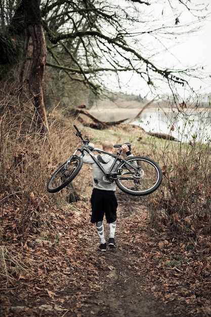 Rear of man carrying bicycle on his back walking near the lake