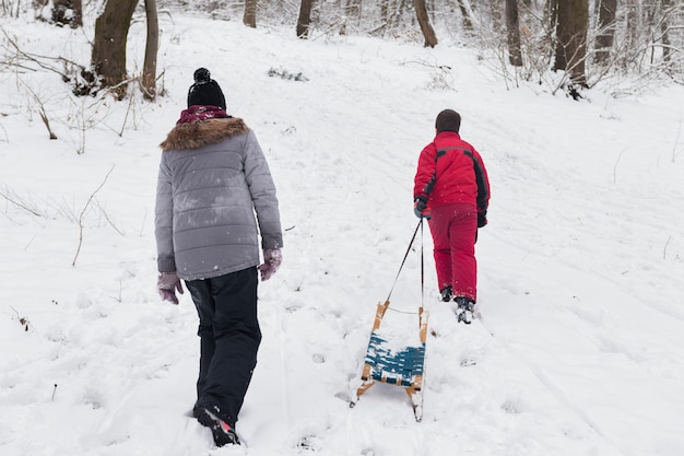 Rear view of boy and girl walking with empty sledge in snowy forest