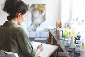 Free photo rear view of brunette young caucasian woman artist in khaki shirt holding pencil, sketching at workshop with paints on table near her. art, creativity, painting, hobby, job and occupation concept