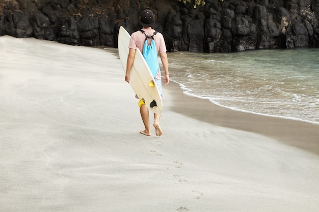 Free photo rear view of caucasian barefooted surfer carrying surfboard while walking along beach with rocky shore, heading to surfing spot