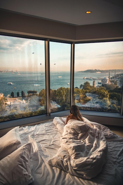 Free photo rear view of a female lying on bed looking at the beach through the glass windows