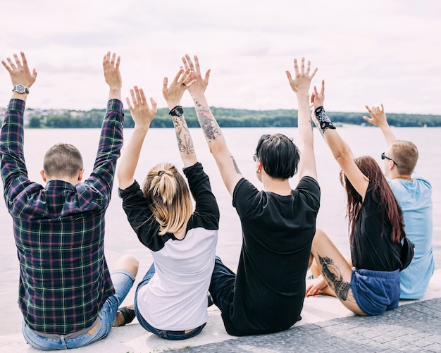 Rear view of friends sitting near the lake raising their hands