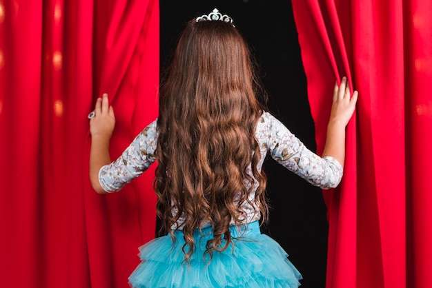 Rear view of girl with long brunette wavy hair looking from red curtain