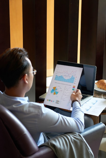 Rear view of man seated in armchair in cafe analyzing diagrams and charts before presentation