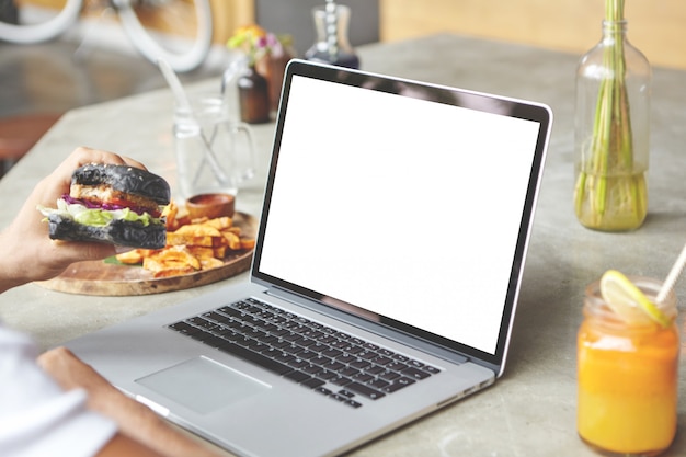 Rear view of student sitting in front of open generic laptop with burger in his hand