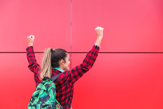 Free Photo rear view of teenage girl carrying bag at back raising their hands against red background