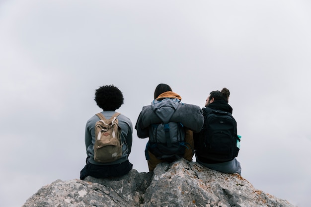 Free photo rear view of a three friends sitting on rock against sky