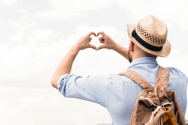 Free photo rear view of traveler man making heart shape from finger against cloudy sky