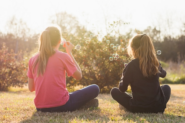 Rear view of two girl sitting on green grass blowing bubbles