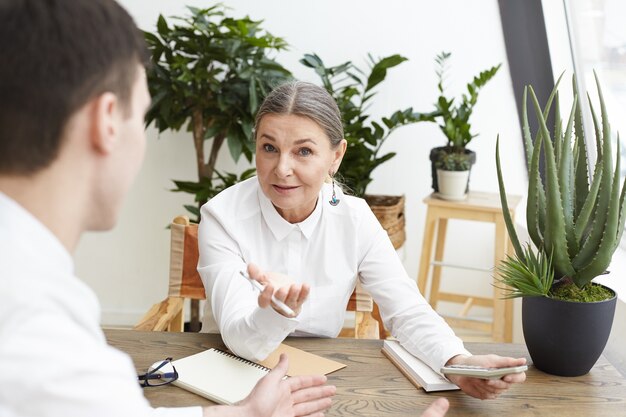 Rear view of unrecognizable young male candidate having job interview with confident middle aged gray haired woman recruiter who is holding pencil and calculator, telling him about desired position