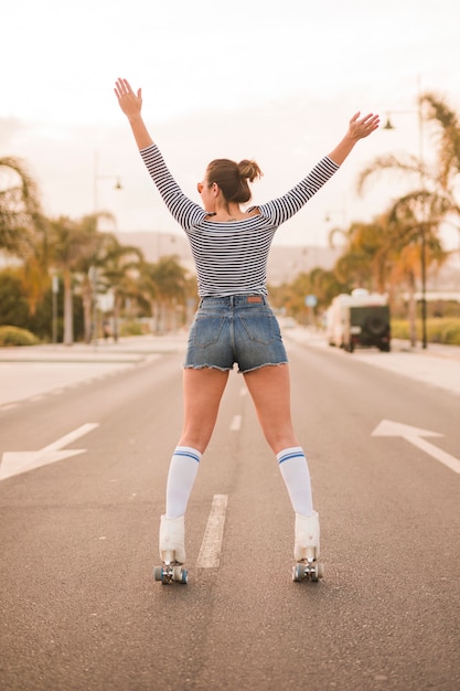 Rear view of a woman standing on road raising her hands balancing on the rolling skate