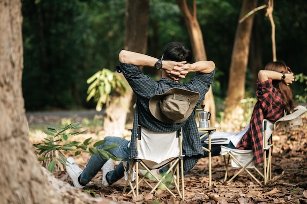 Rear view, Young Asian teenager couple have relax time with camping trip, They are sitting and hands on back of neck on chair in front of backpack camping tent in forest