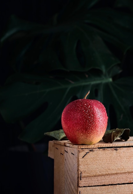 Free photo red apple in water drops on a wooden box on a dark background with monstera leaves closeup selective focus