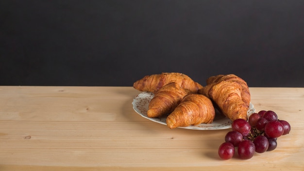 Free photo red bunch of grapes and croissant plate on desk against black background