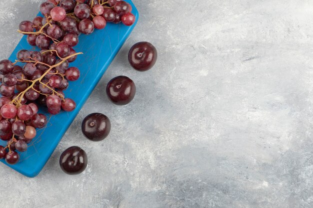 Red grapes on blue cutting board with fresh plums on marble surface.