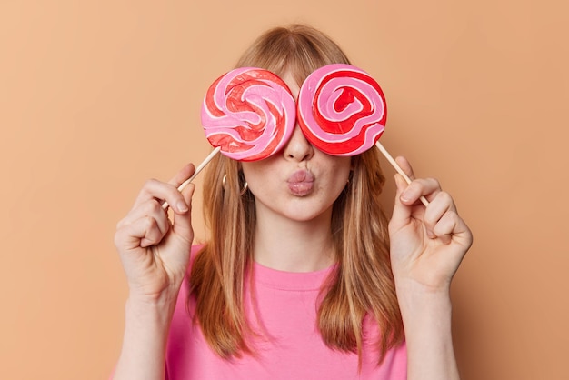 Free photo red haired girl covers eyes with round caramel candies on sticks keeps lips folded has sweet tooth sugar addiction wears casual t shirt isolated over beige background female teenager with lollipops