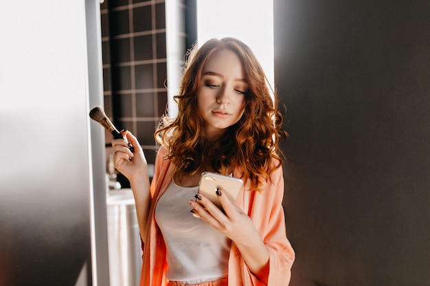 Free photo red-haired girl making makeup in bathroom. photo of serious ginger woman posing with phone at home.