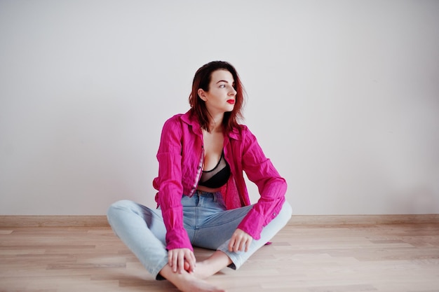 Red haired girl with a big bust on black bra and rose shirt against white wall at empty room