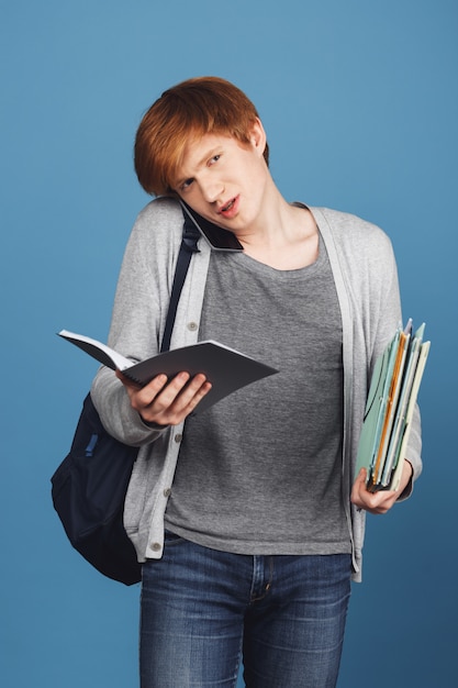 Free Photo red-haired young good-looking male student in casual outfit with black backpack holding lot of books and notebook in hands, talking on phone with mother.