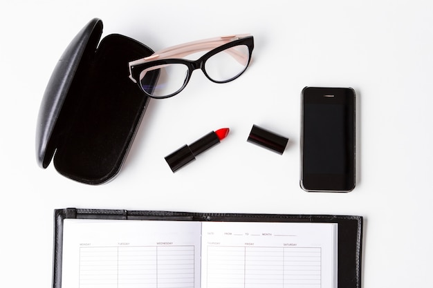 Red lipstick phone glasses and notebook over white surface Above.