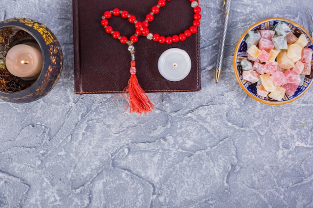 Free photo red prayer beads; candle; diary; pen; lighted candle with bowl of rakhat-lukum on rough grey textured background