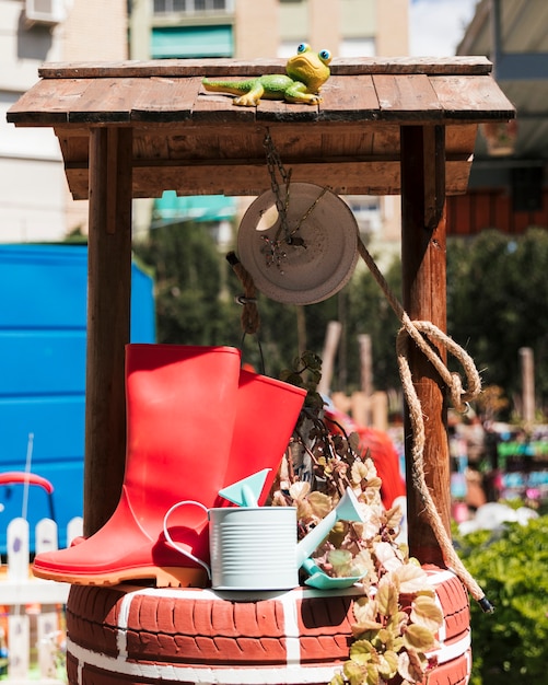 Free Photo red rubber boot; watering can and spade over the handmade well in the garden