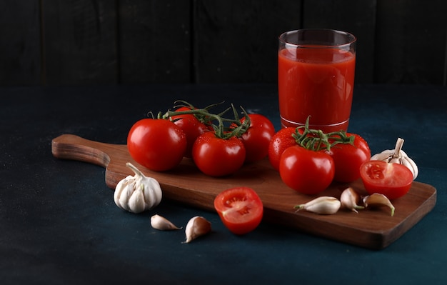 Free photo red tomatoes and garlic gloves on the wooden board with a glass of juice on the blue background.