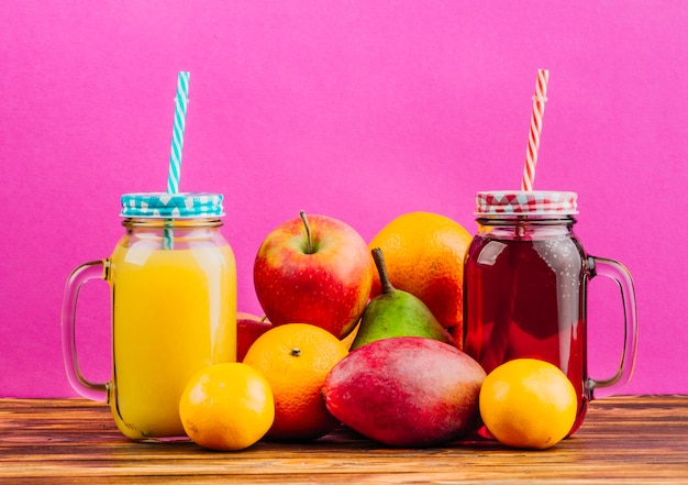 Free photo red and yellow juice mason jars with drinking straws and fresh fruits against pink background