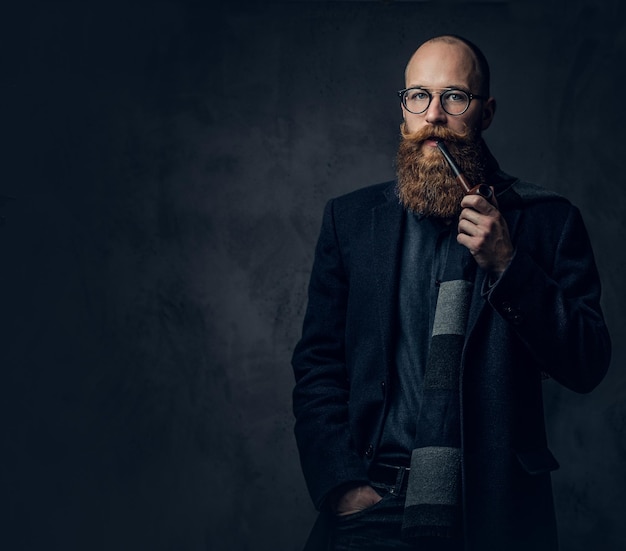 Redhead bearded male dressed in a suit and eyeglasses smoking tradition pipe over dark grey background.