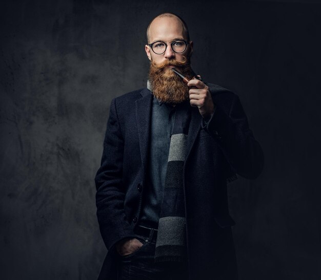 Redhead bearded male dressed in a suit and eyeglasses smoking tradition pipe over dark grey background.