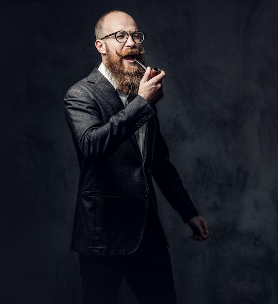 Redhead bearded male dressed in a suit and eyeglasses smoking tradition pipe over dark grey background.