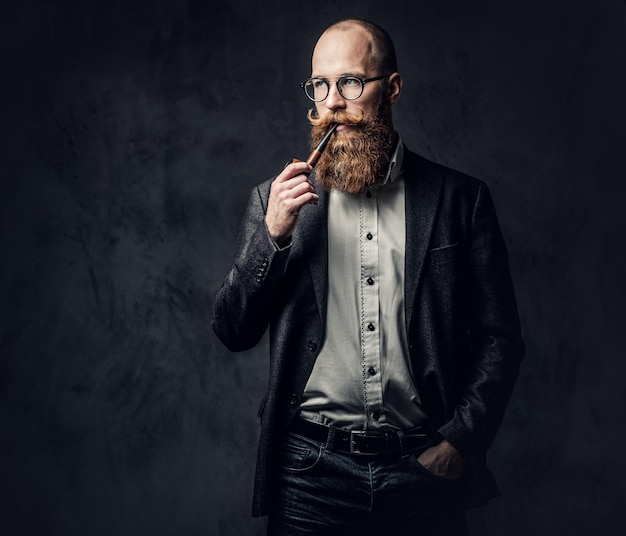 Redhead bearded male dressed in a suit and eyeglasses smoking tradition pipe over dark grey background.